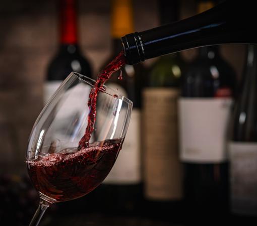 A dark, elegant still life of red wine being poured into a glass with wine bottles and fresh grapes in the background. Perfect for wine-themed compositions